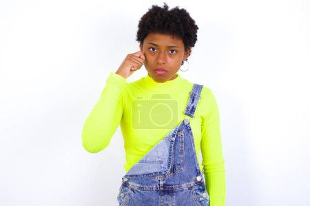 Foto de Mujer afroamericana joven infeliz con el pelo corto usando denim en general contra la pared blanca llorando mientras posando en la cámara batiendo lágrimas con la mano. - Imagen libre de derechos