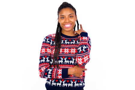 Foto de Mujer afroamericana alegre usando suéter de Navidad contra la pared blanca con la mano cerca de la cara. Mirando con expresión alegre a la cámara después de escuchar buenas noticias. Confianza.. - Imagen libre de derechos
