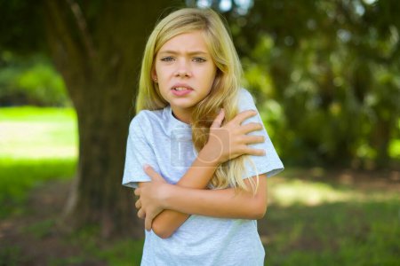 Portrait of caucasian little kid girl wearing white t-shirt standing outdoor in the park hugging herself when feeling cold 