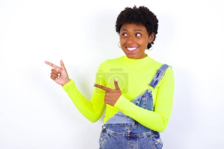 Foto de Joven mujer afroamericana optimista con el pelo corto que usa denim en general contra los puntos blancos de la pared con ambas manos y mirando el espacio vacío. - Imagen libre de derechos