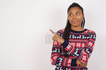 Photo for Portrait of African American woman wearing Christmas sweater against white wall posing on camera with tricky look, presenting product with index finger. Advertisement concept. - Royalty Free Image
