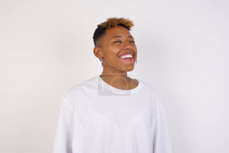 Photo for Joyful african-american woman wearing white t-shirt having fun and laughs at good joke or sings wears casual clothes, standing against gray wall. Happy woman with long hair poses indoors. - Royalty Free Image