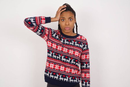 Téléchargez les photos : Portrait de femme afro-américaine confus portant un pull de Noël contre un mur blanc tenant la main sur les cheveux et fronçant les sourcils, paniquant, perdant la mémoire. Inquiète et anxieuse ne se souvient de rien. - en image libre de droit