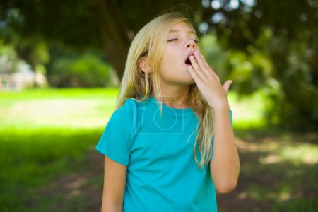 Photo for Sad caucasian little kid girl wearing blue t-shirt standing outdoor in the park feeling sleepy - Royalty Free Image