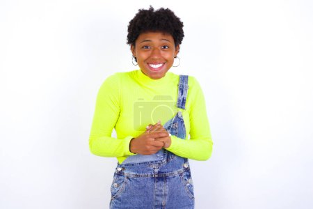 Photo for Happy young African American woman with short hair wearing denim overall against white stands against orange studio wall keeps hands on heart, swears be loyal, expresses gratitude. Honesty concept. - Royalty Free Image