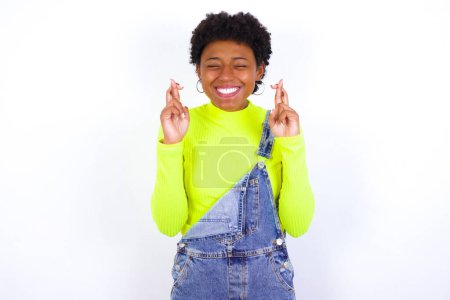 Foto de Joven mujer afroamericana alegre con el pelo corto que usa denim en general contra la pared blanca aprieta los dientes, levanta los dedos cruzados, hace el deseo deseable, espera buenas noticias, tengo que ganar. - Imagen libre de derechos