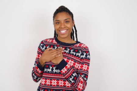 Foto de Honesta mujer afroamericana vistiendo suéter de Navidad contra la pared blanca mantiene las manos en el pecho, tocado por el cumplido o hace la promesa, mira a la cámara con gran placer. - Imagen libre de derechos