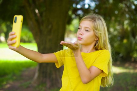 Téléchargez les photos : Petite fille caucasienne portant t-shirt jaune debout à l'extérieur souffle baiser d'air à la caméra du smartphone et prend selfie, envoie mwah via appel en ligne. - en image libre de droit