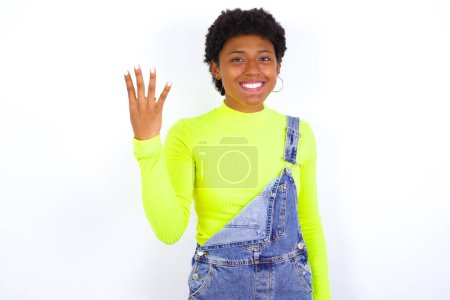 Foto de Joven mujer afroamericana con el pelo corto con denim en general contra la pared blanca sonriendo y mirando amigable, mostrando el número cuatro o cuarto con la mano hacia adelante, cuenta atrás - Imagen libre de derechos