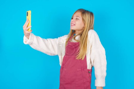 Isolated shot of pleased cheerful pretty teen girl , makes selfie with mobile phone. People, technology and leisure concept