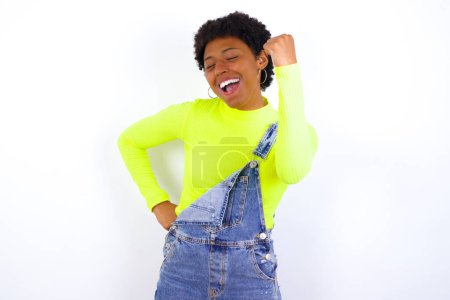 Photo for Overjoyed young African American woman with short hair wearing denim overall against white glad to receive good news, clenching fist and making winning gesture. - Royalty Free Image
