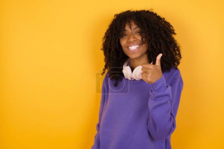 Téléchargez les photos : Portrait de jeune femme afro-américaine expressive avec écouteurs sur fond jaune montrant pouce vers le haut - en image libre de droit