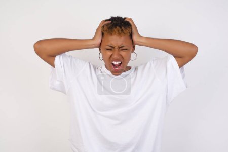 Photo for Shocked panic young African American woman wearing white t-shirt holding hands on head and screaming in despair and frustration while being late for plane, her eyes full of terror, mouth dropped open - Royalty Free Image