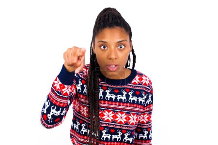 Foto de Mujer afroamericana impactada vistiendo suéter de Navidad contra los puntos blancos de la pared frontal con el dedo índice en la cámara y. Concepto de sorpresa y publicidad. - Imagen libre de derechos