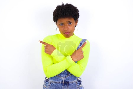 Photo for Serious young African American woman with short hair wearing denim overall against white crosses hands and points at different sides hesitates between two items. Hard decision concept - Royalty Free Image