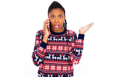 Téléchargez les photos : Femme afro-américaine portant un pull de Noël contre un mur blanc parlant au téléphone stressé avec la main sur le visage, choqué par la honte et le visage surprise, en colère et frustré. Peur et contrariété par erreur. - en image libre de droit
