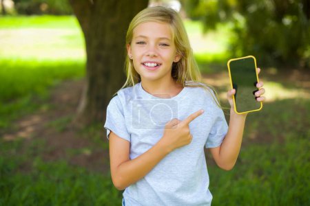 Photo for Portrait of caucasian little kid girl wearing white t-shirt standing outdoor in the park pointing finger on mobile phone - Royalty Free Image