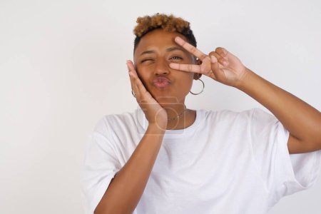 Photo for Leisure lifestyle people celebrate flirt coquettish concept. Beautiful pretty cheerful gorgeous young African American woman wearing white t-shirt showing making v-sign near eyes wearing casual clothes standing against gray wall. - Royalty Free Image