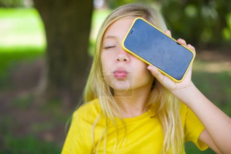 Foto de Adorable niña caucásica vistiendo camiseta amarilla de pie al aire libre sosteniendo dispositivo moderno que cubre el ojo con los labios enfurecidos - Imagen libre de derechos