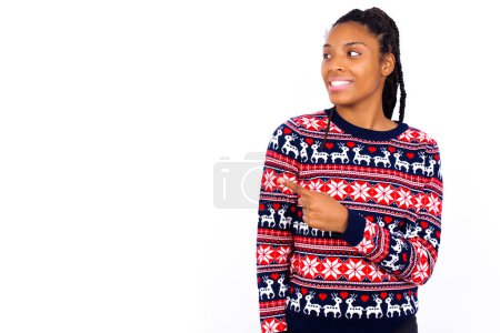 Téléchargez les photos : Joyeux sourire femme afro-américaine souriante portant un pull de Noël contre un mur blanc regardant et pointant de côté avec la main. Espace de copie et concept publicitaire. - en image libre de droit