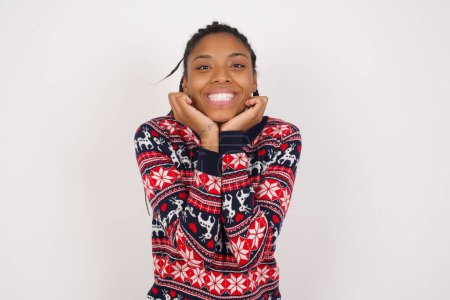 Téléchargez les photos : Femme afro-américaine rêveuse portant un pull de Noël contre un mur blanc garde les mains serrées sous le menton, regarde avec une expression heureuse, a sourire aux dents. - en image libre de droit