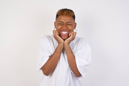 Portrait of young cute black student being overwhelmed with emotions, expressing excitement and happiness with closed eyes and hands near face while smiling broadly over gray background.