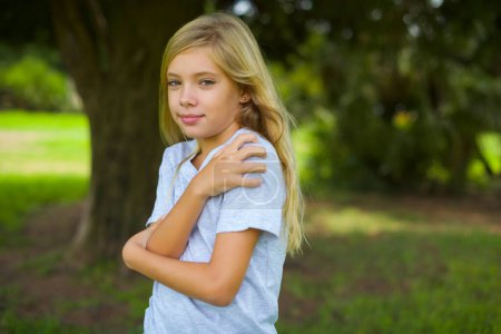 Photo for Charming pleased caucasian little kid girl wearing white t-shirt standing outdoor in the park embraces own body, pleasantly feels comfortable poses. Tenderness and self esteem concept - Royalty Free Image