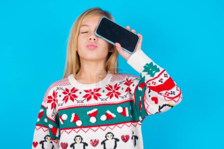 Téléchargez les photos : Adorable petite fille caucasienne portant un pull de Noël tricoté sur fond bleu tenant un dispositif moderne couvrant les yeux avec des lèvres boudées - en image libre de droit
