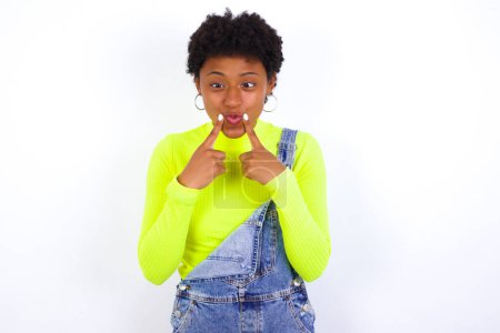 Téléchargez les photos : Jeune femme afro-américaine aux cheveux courts portant le denim dans l'ensemble contre le mur blanc traverse les yeux et rend les lèvres de poisson drôle grimace - en image libre de droit