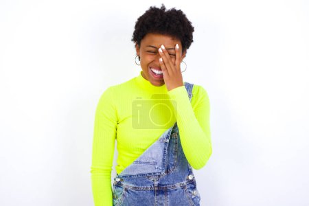 Téléchargez les photos : Jeune femme afro-américaine avec les cheveux courts portant le denim dans l'ensemble contre le blanc rend la paume du visage et sourit largement, ricane entend positivement pose drôle de blague - en image libre de droit