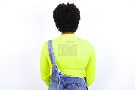 Photo for Young African American woman with short hair wearing denim overall against white standing backwards looking away with arms on body. - Royalty Free Image