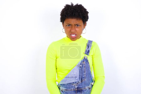 Photo for Mad crazy young African American woman with short hair wearing denim overall against white wall clenches teeth angrily, being annoyed with coming noise. Negative feeling concept. - Royalty Free Image
