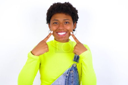 Foto de Fuertes dientes blancos rectos sanos. Retrato de cerca de la joven mujer afroamericana feliz con el pelo corto usando denim en general contra la pared blanca con sonrisa radiante apuntando a los dientes blancos claros perfectos. - Imagen libre de derechos