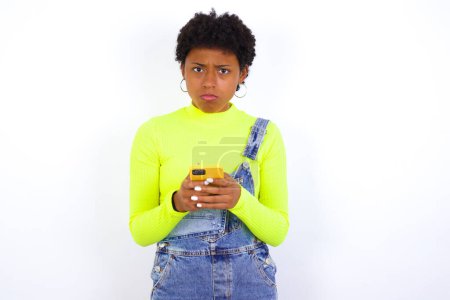 Foto de Retrato de una joven afroamericana confusa con el pelo corto usando denim en general contra la pared blanca sosteniendo el teléfono móvil y encogiéndose de hombros y frunciendo el ceño. - Imagen libre de derechos