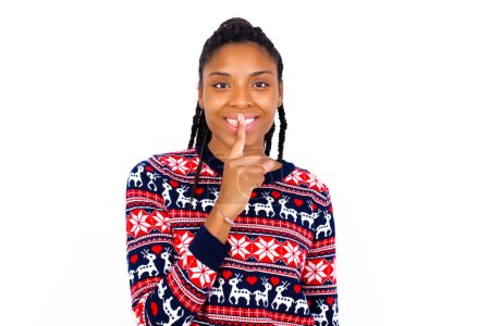 Foto de Sonriente mujer afroamericana vistiendo suéter de Navidad contra la pared blanca hace gesto de silencio, sostiene el dedo delantero sobre los labios se esconde secreto. Silencio, por favor.. - Imagen libre de derechos