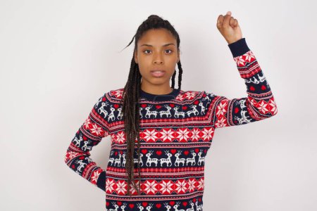 Téléchargez les photos : Femme afro-américaine portant un pull de Noël contre un mur blanc se sentant sérieuse, forte et rebelle, levant le poing, protestant ou se battant pour la révolution. - en image libre de droit