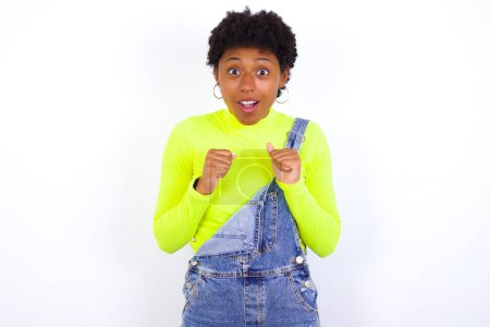 Photo for Portrait of desperate and shocked young African American woman with short hair wearing denim overall against white wall looking panic, holding hands near face, with mouth wide open. - Royalty Free Image