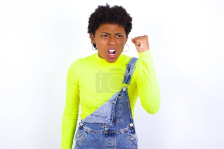 Téléchargez les photos : Jeune femme afro-américaine féroce avec les cheveux courts portant le denim dans l'ensemble contre le poing blanc tenant devant comme si elle était prête pour le combat ou le défi, criant et ayant une expression agressive sur le visage. - en image libre de droit