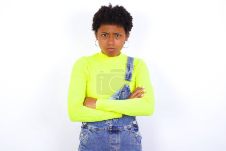 Photo for Gloomy dissatisfied young African American woman with short hair wearing denim overall against white looks with miserable expression at camera from under forehead, makes unhappy grimace - Royalty Free Image