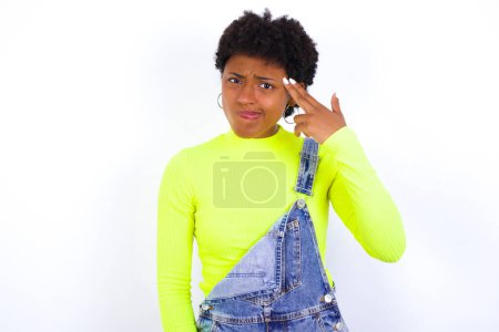 Foto de Mujer afroamericana infeliz joven con el pelo corto con denim en general contra la pared blanca imita disparo de arma hace gesto suicida mantiene dos dedos en las sienes. - Imagen libre de derechos