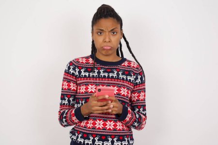 Photo for Portrait of a confused African American woman wearing Christmas sweater against white wall holding mobile phone and shrugging shoulders and frowning face. - Royalty Free Image