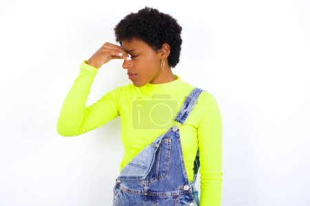 Téléchargez les photos : Triste jeune afro-américaine aux cheveux courts portant globalement du denim contre un mur blanc souffrant de maux de tête se tenant la main sur le visage - en image libre de droit