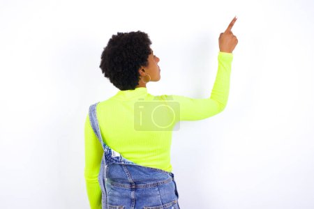 Foto de Joven mujer afroamericana con el pelo corto con denim en general contra la pared blanca que apunta a objeto en el espacio de copia, vista trasera. Da la espalda. - Imagen libre de derechos