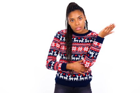 Téléchargez les photos : Studio shot of frustrated African American woman wearing Christmas sweater against white wall gesturing with raised palm, frowning, being displeased and confused with dumb question. - en image libre de droit