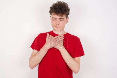 Photo for Handsome young man over white background closes eyes and keeps hands on chest near heart, expresses sincere emotions, being kind hearted and honest. Body language and real feelings concept. - Royalty Free Image
