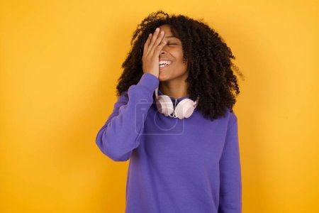 Foto de Retrato de joven mujer afroamericana expresiva con auriculares sobre fondo amarillo riendo y cubriendo la cara - Imagen libre de derechos