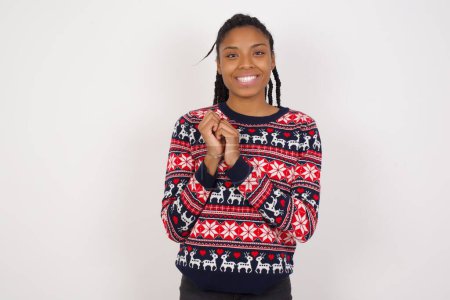 Foto de Mujer afroamericana encantadora soñadora que usa suéter de Navidad contra la pared blanca con expresión agradable, mantiene las manos cruzadas cerca de la cara, emocionadas por algo agradable. - Imagen libre de derechos