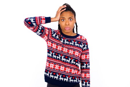 Foto de Retrato de una mujer afroamericana confusa usando suéter navideño contra una pared blanca sosteniendo el pelo y frunciendo el ceño, entrando en pánico, perdiendo la memoria. Preocupado y ansioso no puede recordar nada. - Imagen libre de derechos