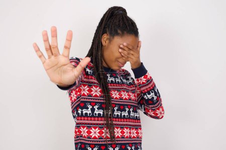 Foto de Mujer afroamericana vistiendo suéter de Navidad contra la pared blanca cubre los ojos con la palma y haciendo gesto de parada, trata de esconderse. No me mires, no quiero ver, me da vergüenza o miedo. - Imagen libre de derechos
