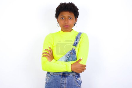 Foto de Joven mujer afroamericana confiada con el pelo corto usando denim en general contra la pared blanca con los brazos cruzados mirando a la cámara - Imagen libre de derechos